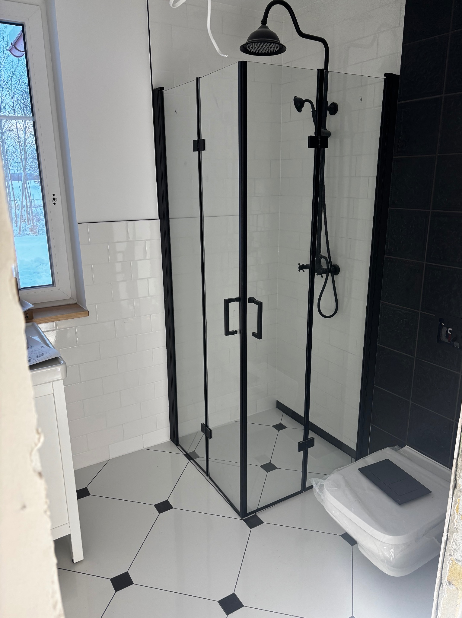 Nowoczesna kabina prysznicowa z czarnymi profilami i deszczownicą w łazience z białymi płytkami i geometryczną podłogą, widoczna częściowo zakryta toaleta w folii ochronnej.