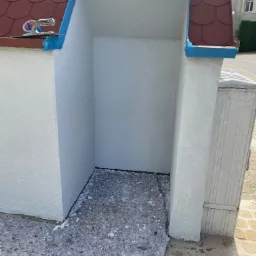 Wnęka zewnętrzna budynku po świeżym pomalowaniu na biało, zabezpieczona niebieską taśmą malarską, z rozłożonym materiałem ochronnym na podłodze i narzędziem do nakładania gładzi na dachu.