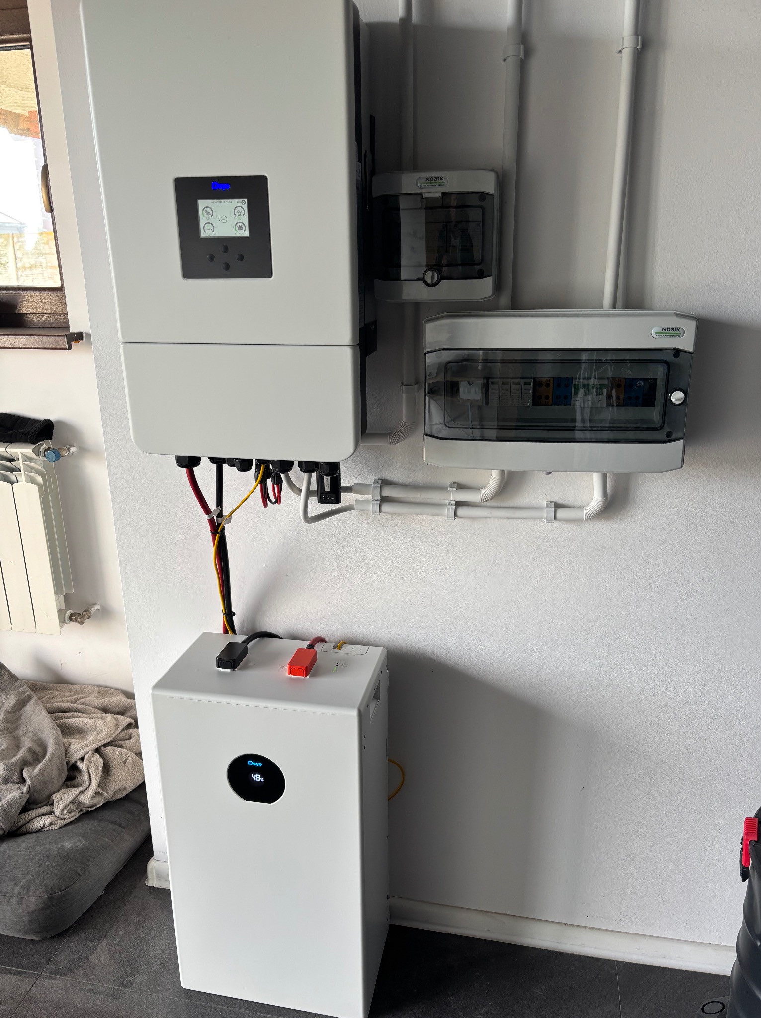 Biały inwerter fotowoltaiczny i magazyn energii Deye na ścianie, z widocznymi przewodami i rozdzielnicami. Instalacja w pomieszczeniu, widoczny fragment okna i grzejnika.