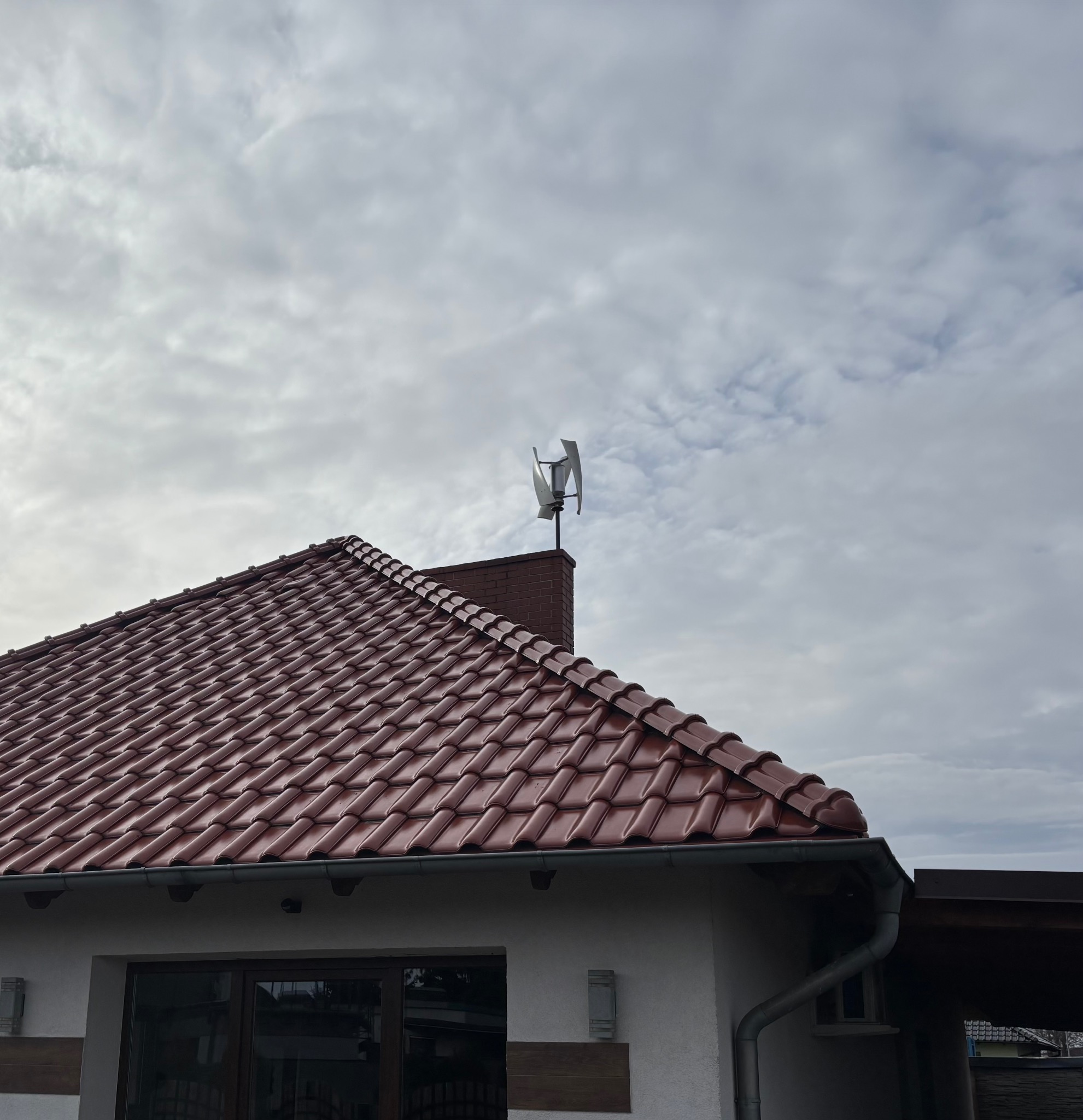 Dom z czerwoną dachówką i turbiną wiatrową na kominie. Pochmurne niebo w tle. Widok z dołu, podkreślający instalację OZE.