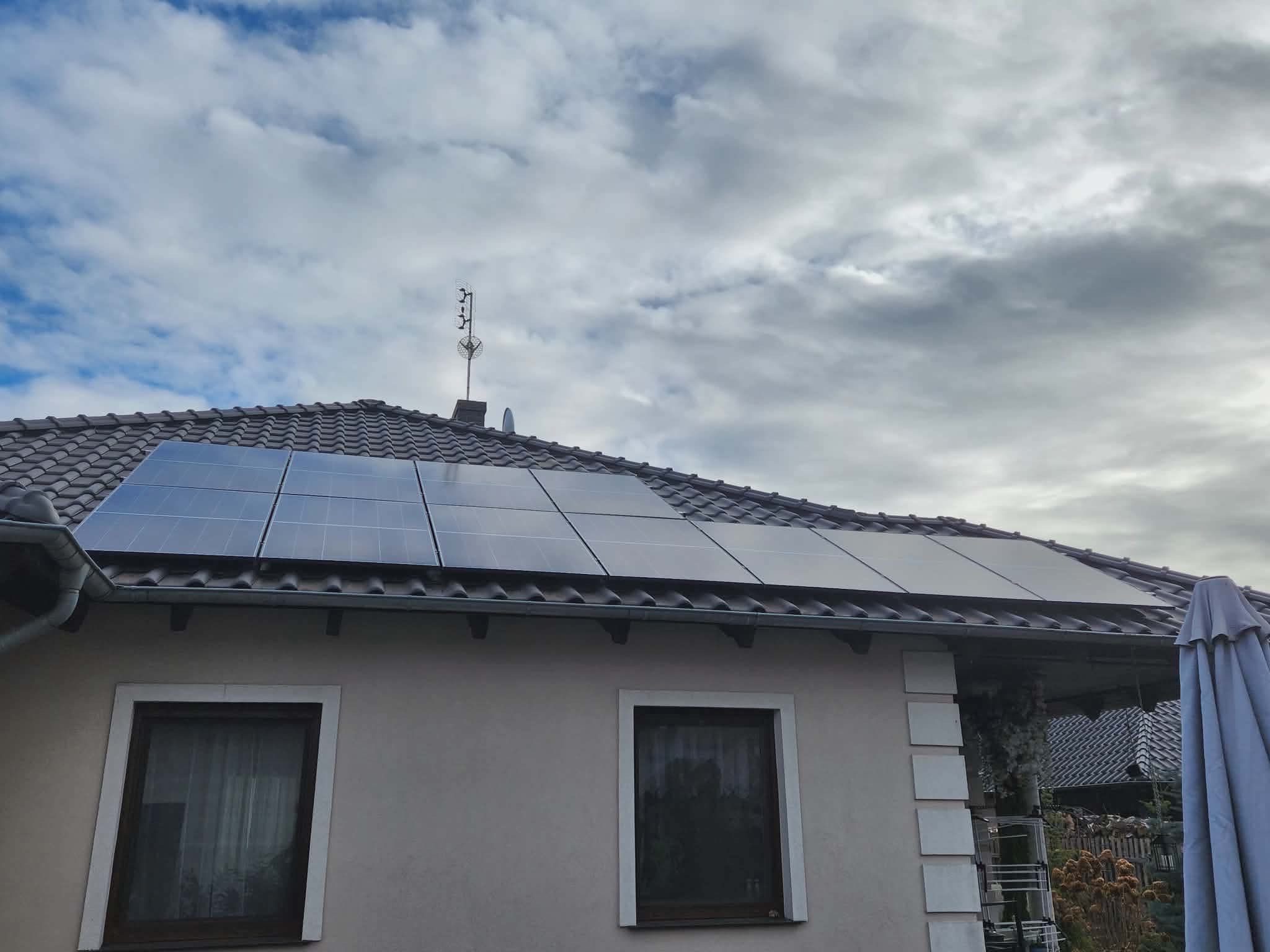Panele fotowoltaiczne na dachu domu jednorodzinnego, widoczne okna i fragment parasola. Pochmurne niebo w tle. Ujęcie z dołu, lekko z boku.