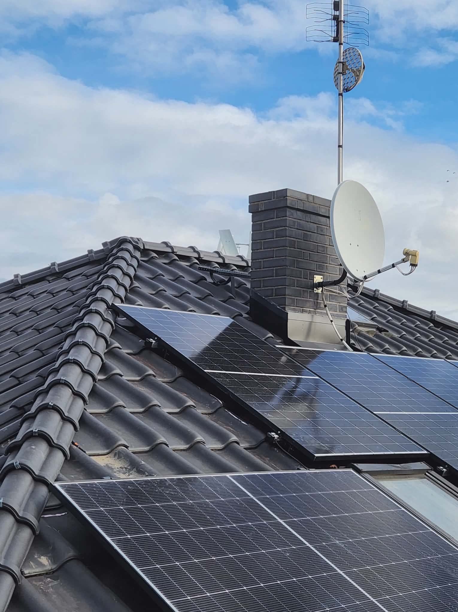 Panele fotowoltaiczne na dachu z czarnej dachówki, komin z cegły, antena satelitarna i telewizyjna na tle błękitnego nieba z chmurami. Widok z góry.