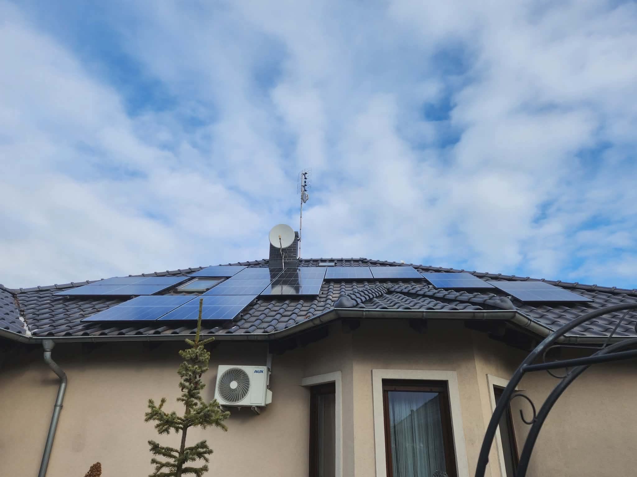 Panele fotowoltaiczne na dachu domu z klimatyzatorem na ścianie. Widoczne okna dachowe i antena satelitarna. Błękitne niebo z chmurami w tle. Ujęcie z dołu.