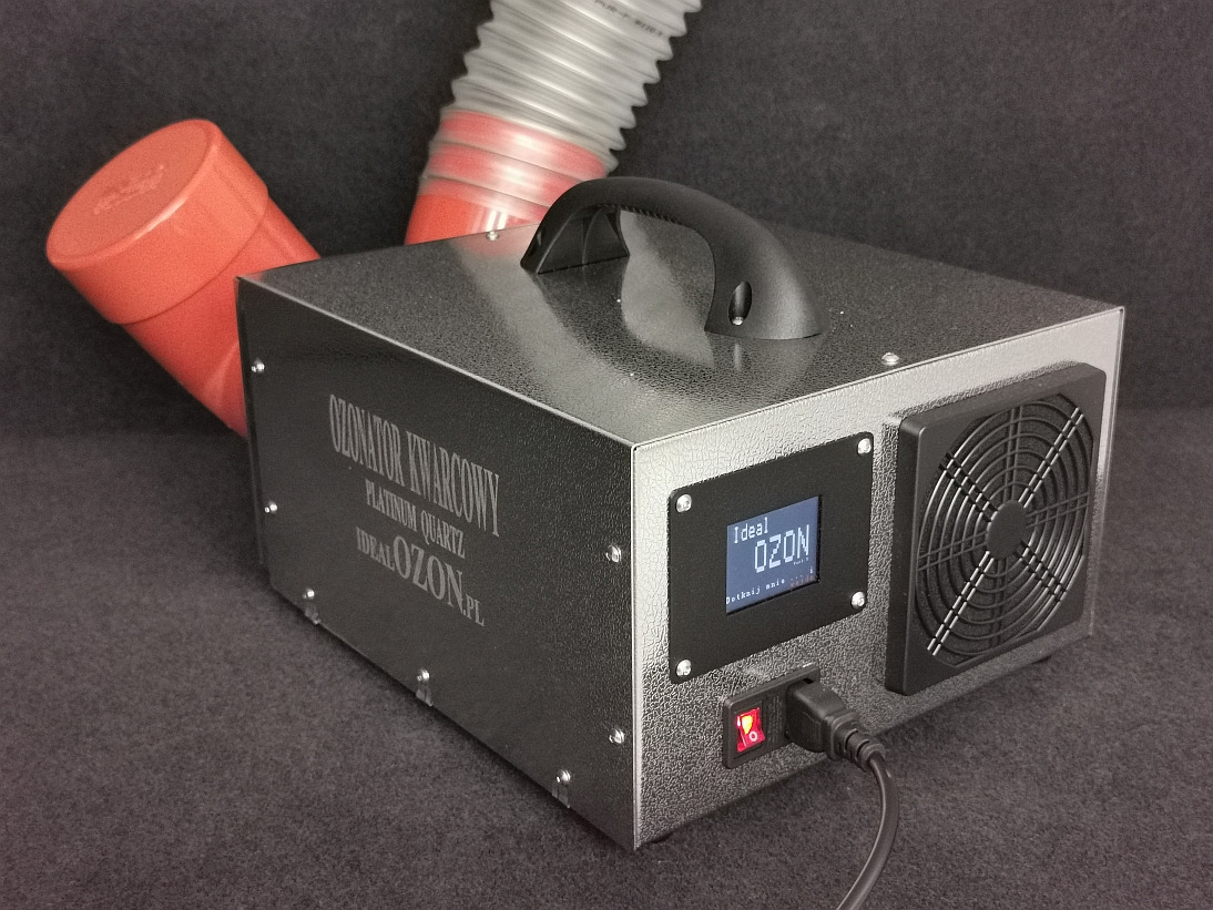 Ozonator kwarcowy w obudowie z fakturą, z wyświetlaczem LCD pokazującym 'Ideal OZON', wentylatorem i podłączonym kablem zasilającym, ustawiony na ciemnoszarym tle z fragmentami czerwonej i szarej...