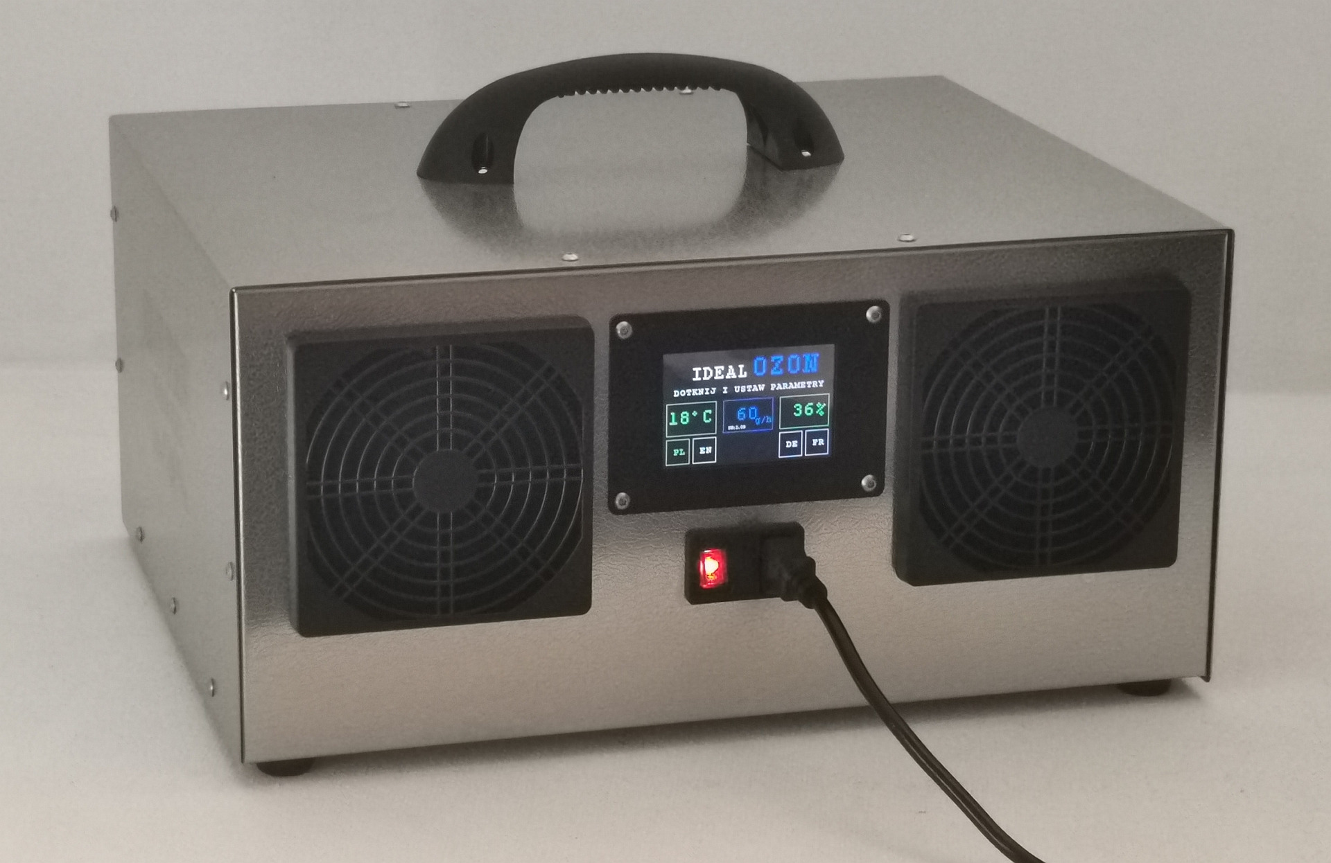 Generator ozonu w metalowej obudowie z czarnym uchwytem i wyświetlaczem LCD pokazującym temperaturę i wilgotność, podłączony do prądu, z widocznymi dwoma wentylatorami.