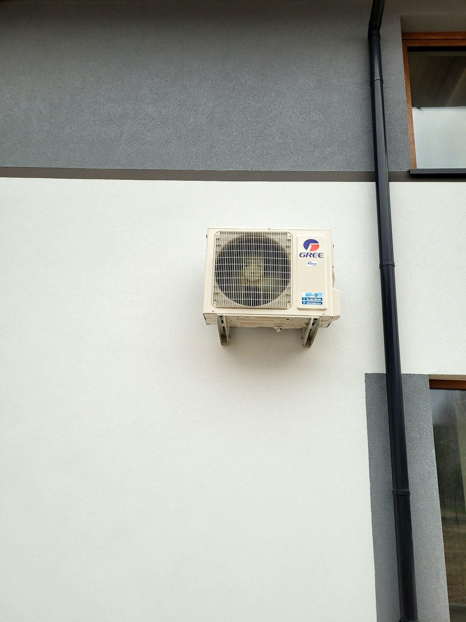 Zamontowana jednostka zewnętrzna klimatyzacji Gree na elewacji budynku z widoczną rurą spustową i oknem.