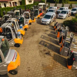 Widok z góry na plac z równo zaparkowanymi wózkami widłowymi marki STILL, w tle budynki i zieleń.