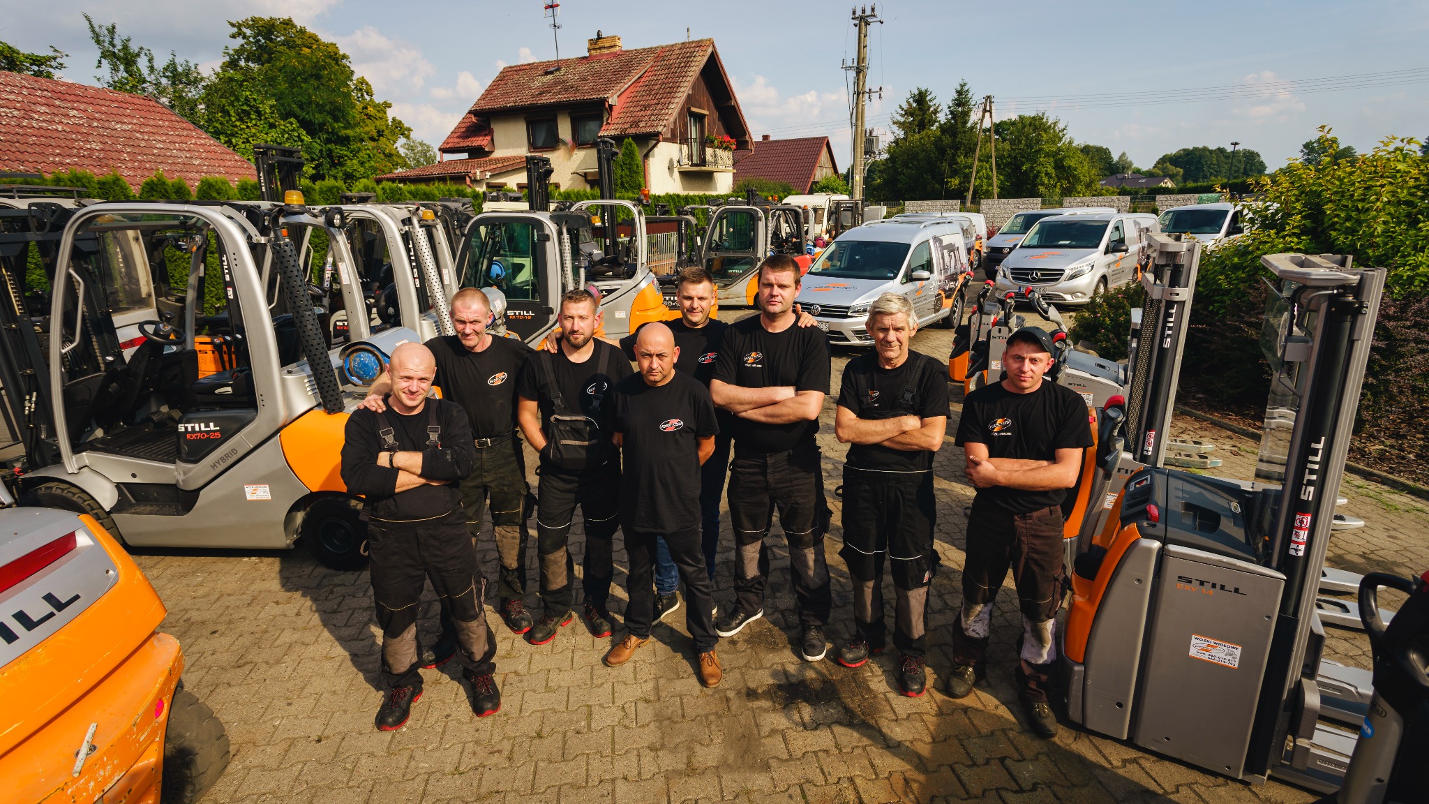 Grupa pracowników w czarnych uniformach stoi przed flotą wózków widłowych marki Still i samochodami dostawczymi na tle budynku mieszkalnego i zieleni.