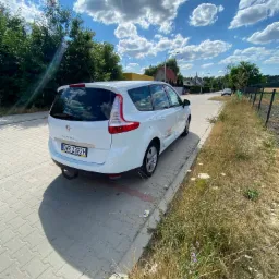 Biały Renault Scenic zaparkowany na chodniku z kostki brukowej, widok z tyłu, tablica rejestracyjna DWR 2392H, hak holowniczy, w tle błękitne niebo z obłokami i zabudowania.