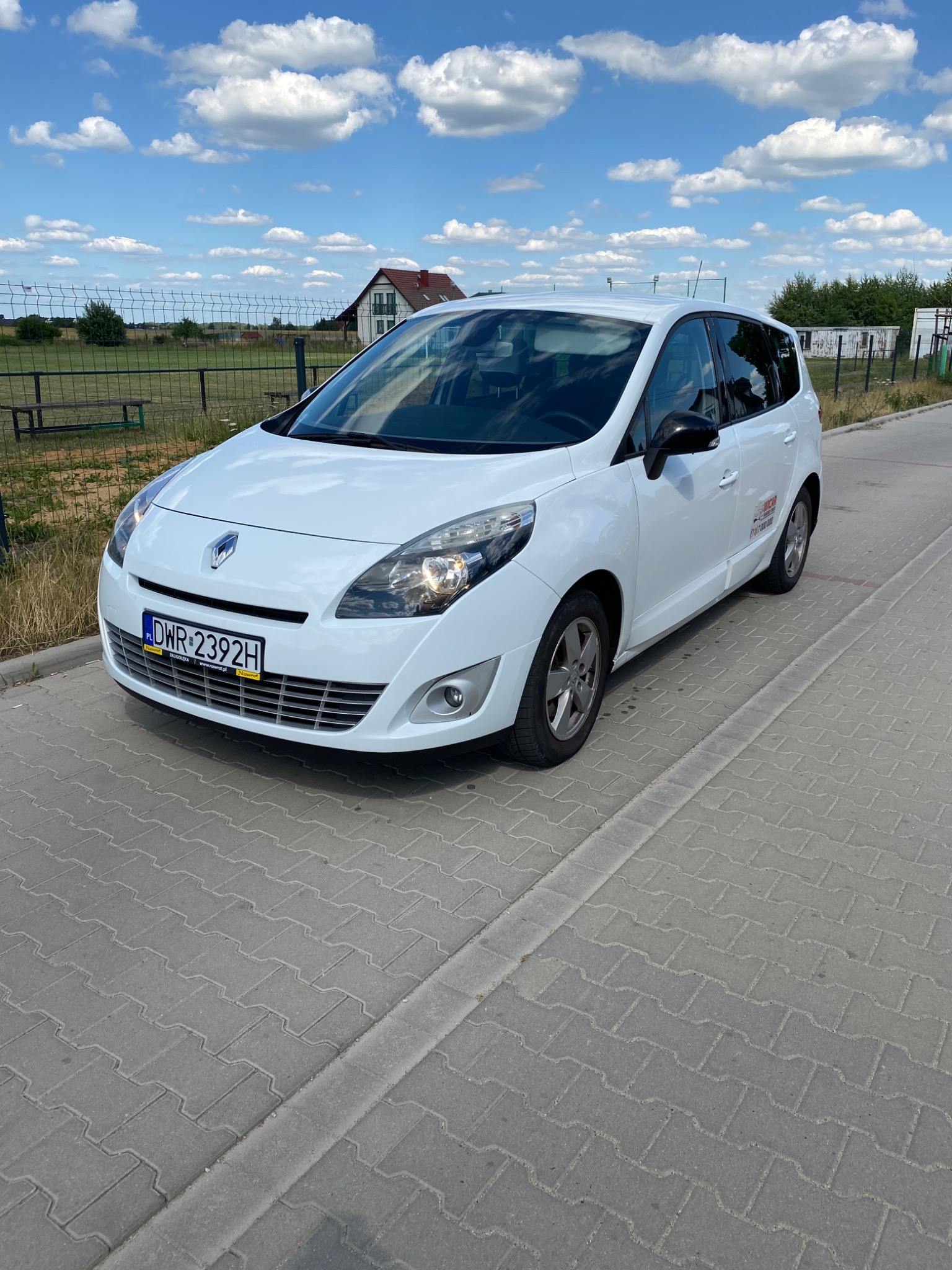 Biały Renault Scenic parkuje na szarej kostce brukowej z widoczną naklejką firmy transportowej na tylnych drzwiach, tło stanowi ogrodzenie i dom.