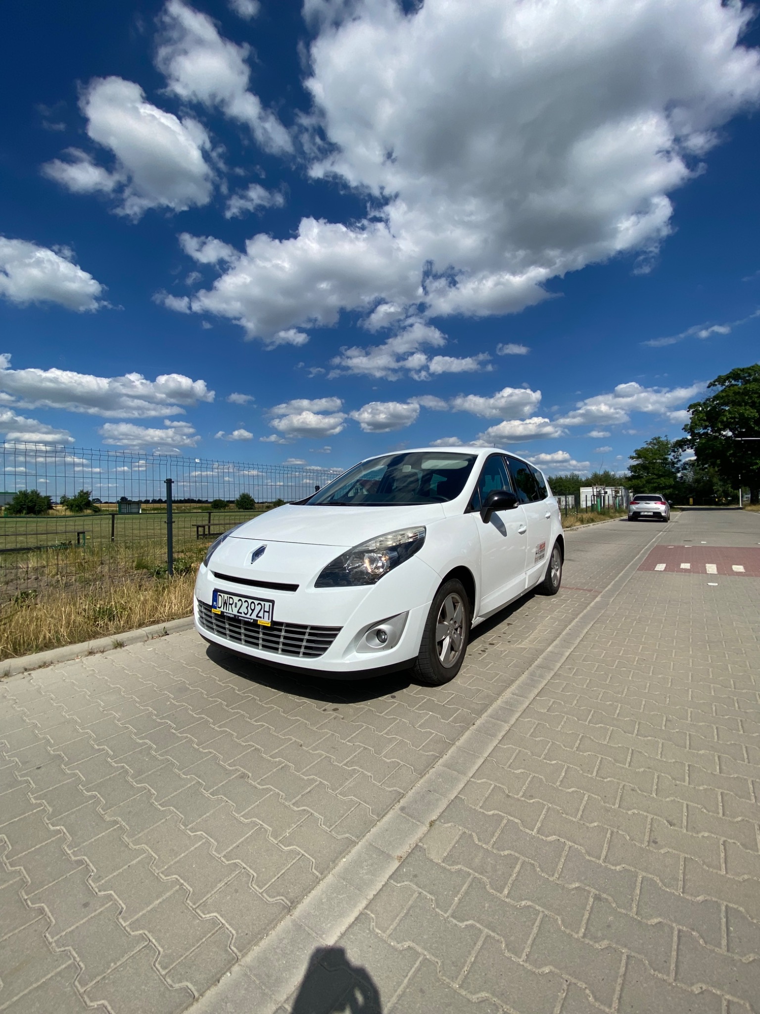 Biały samochód osobowy Renault Scenic zaparkowany na brukowanej drodze w słoneczny dzień z błękitnym niebem i białymi chmurami w tle.