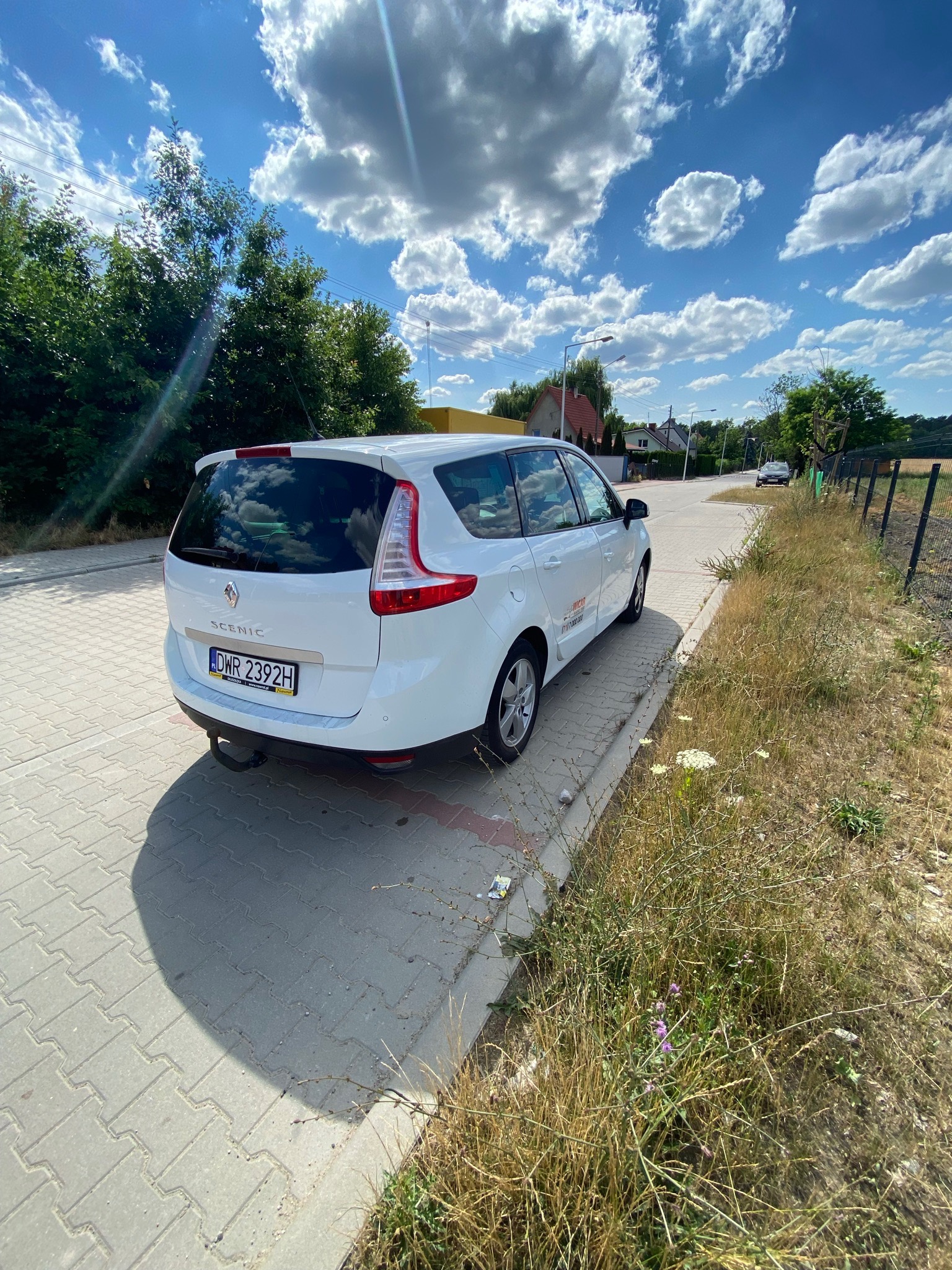 Biały Renault Scenic zaparkowany na chodniku z kostki brukowej, widok z tyłu, tablica rejestracyjna DWR 2392H, hak holowniczy, w tle błękitne niebo z obłokami i zabudowania.