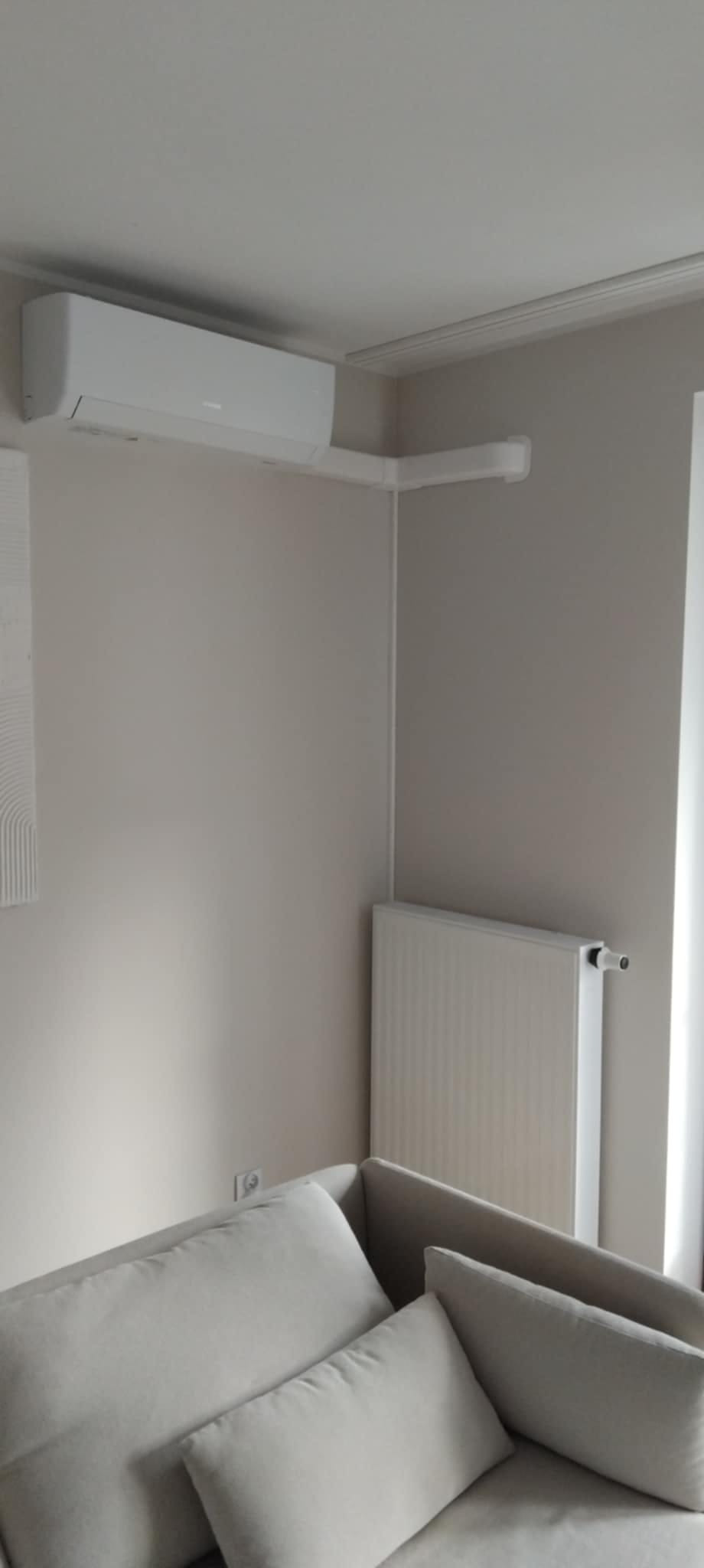 Biały klimatyzator ścienny zamontowany w rogu pokoju nad grzejnikiem, widok z dołu na jasną sofę.