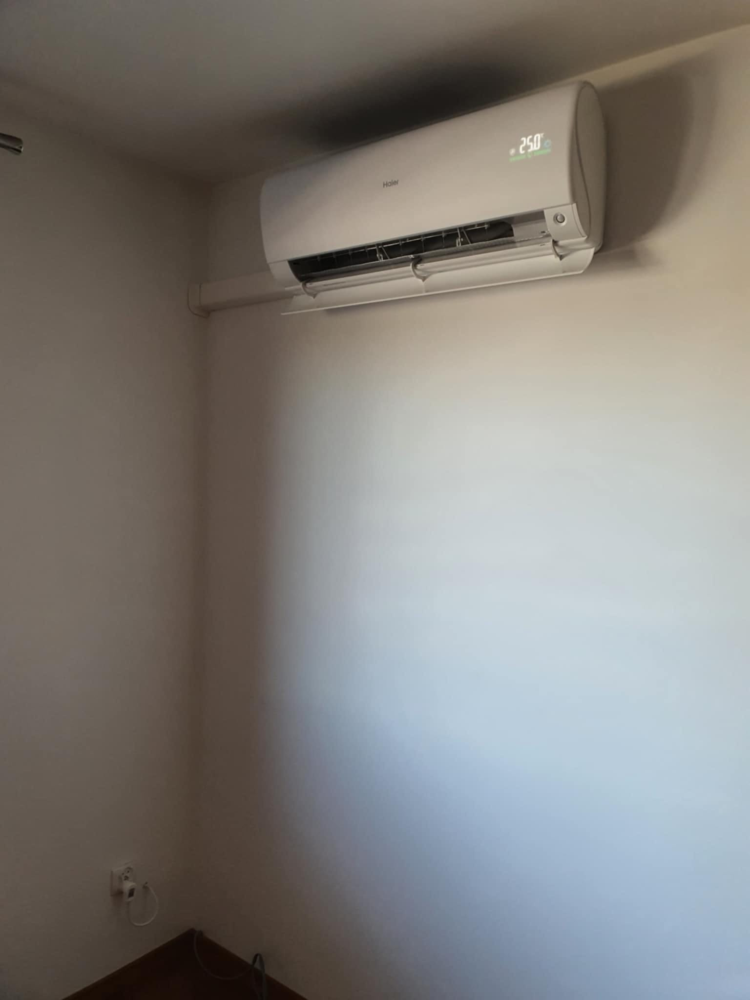 Biały klimatyzator Haier zamontowany na jasnej ścianie w pokoju, wyświetlający temperaturę 25 stopni Celsjusza.