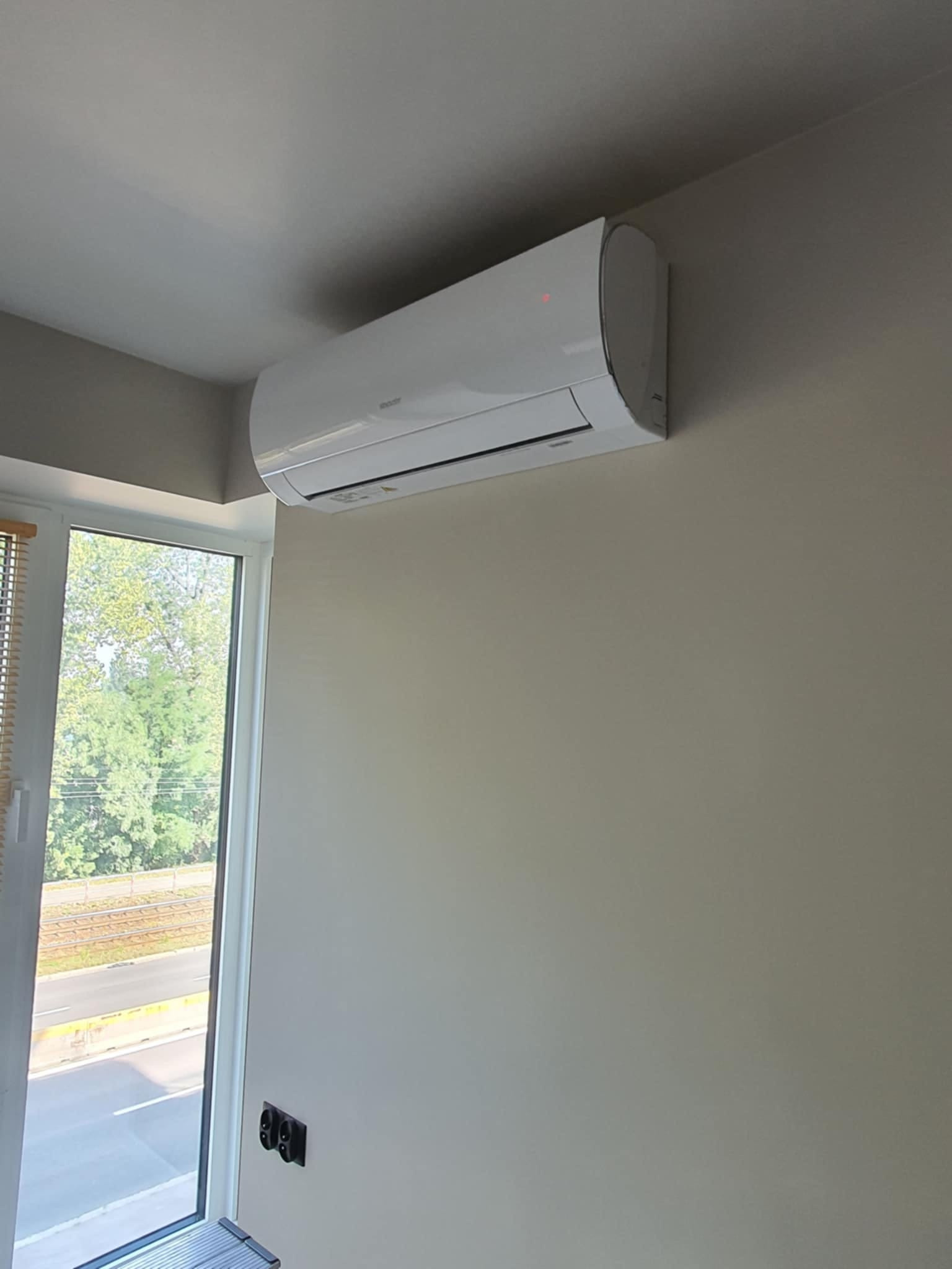 Biały klimatyzator ścienny zamontowany w jasnym pomieszczeniu, widok z boku na urządzenie i okno z widokiem na zieleń.