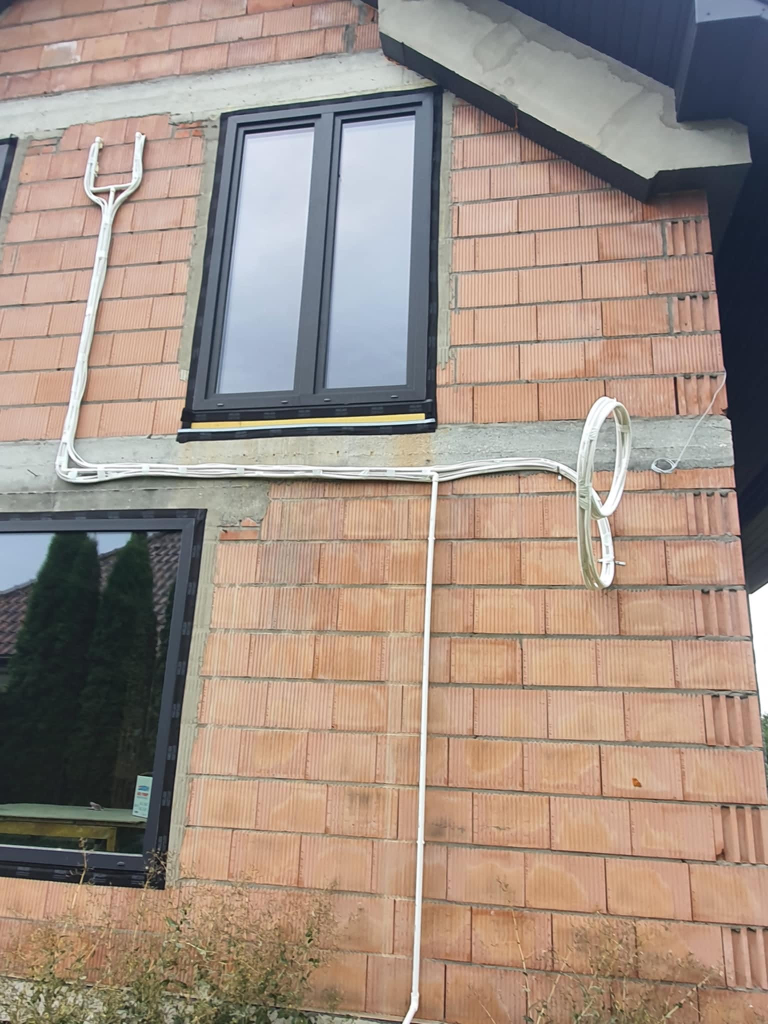 Fragment ściany budynku z pustaków ceramicznych z zamontowanymi oknami w czarnych ramach i poprowadzonymi po elewacji białymi przewodami elektrycznymi w peszlu.