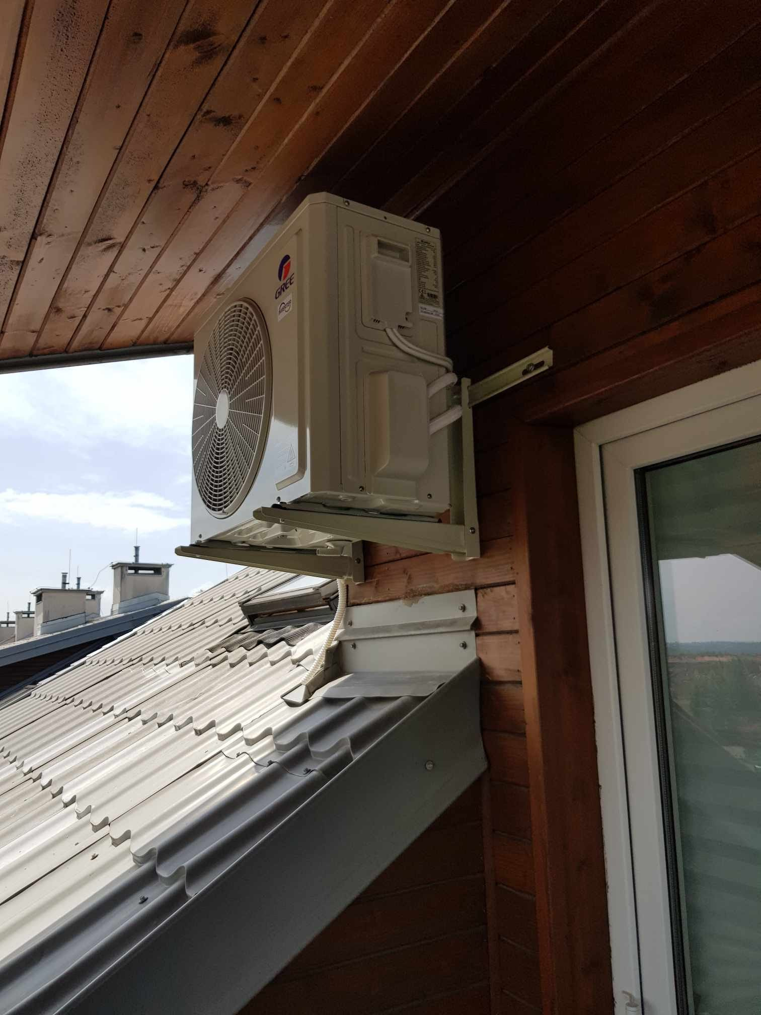 Zewnętrzna jednostka klimatyzacji marki Gree zamontowana na drewnianej elewacji domu, widok z dołu, w tle dach i okno.