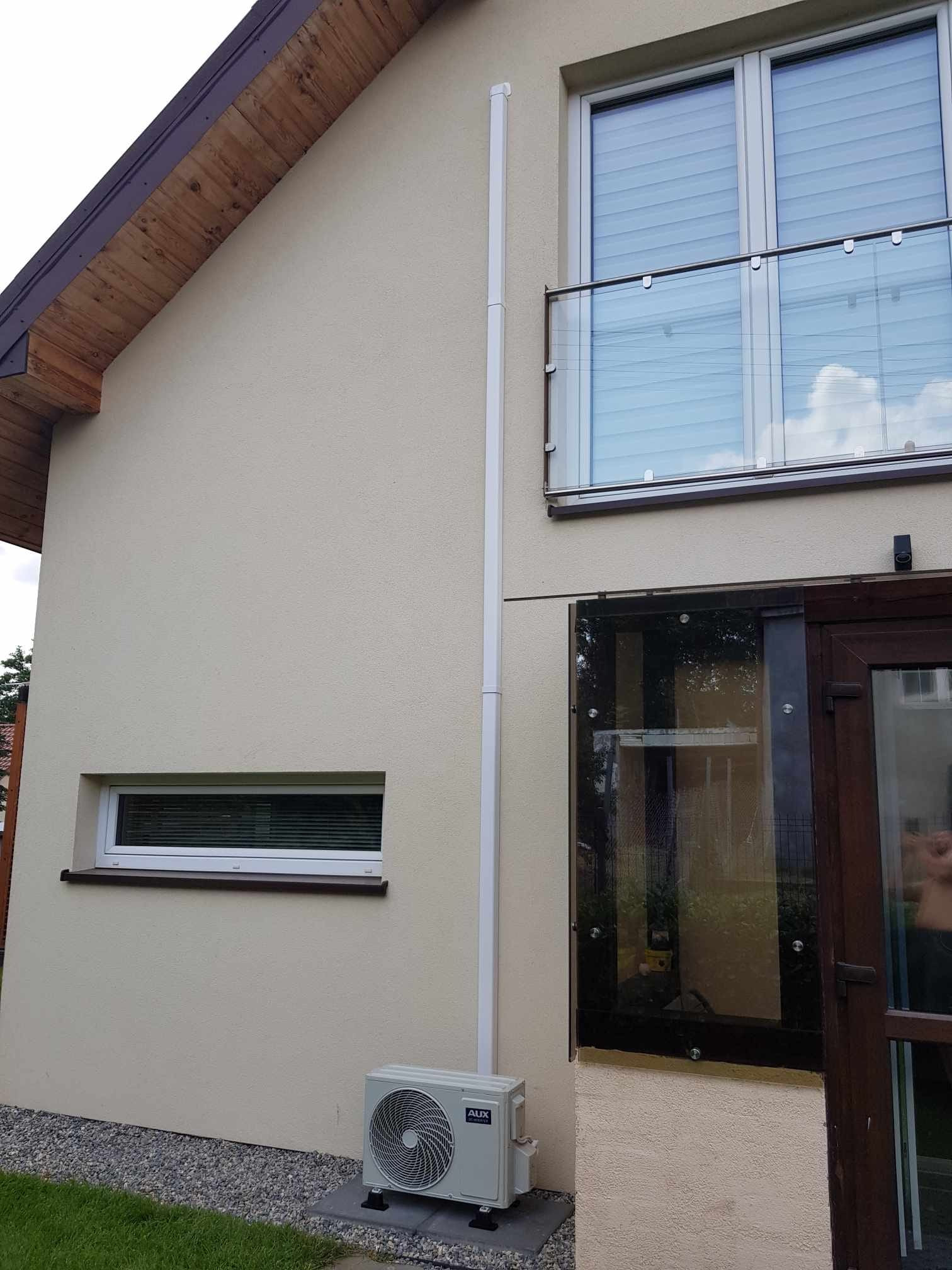 Zewnętrzna jednostka klimatyzacji AUX zamontowana przy ścianie domu z beżową elewacją i grafitowym oknem, widoczna rura odprowadzająca skropliny, na podłożu szary żwir i trawa.