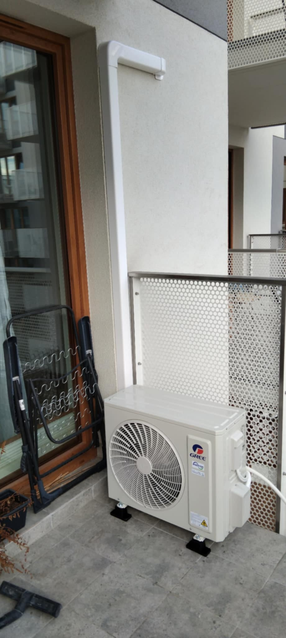 Zewnętrzna jednostka klimatyzacji Gree, zamontowana na balkonie bloku mieszkalnego, widoczna rura odprowadzająca skropliny i złożony suszarka na ubrania obok okna.