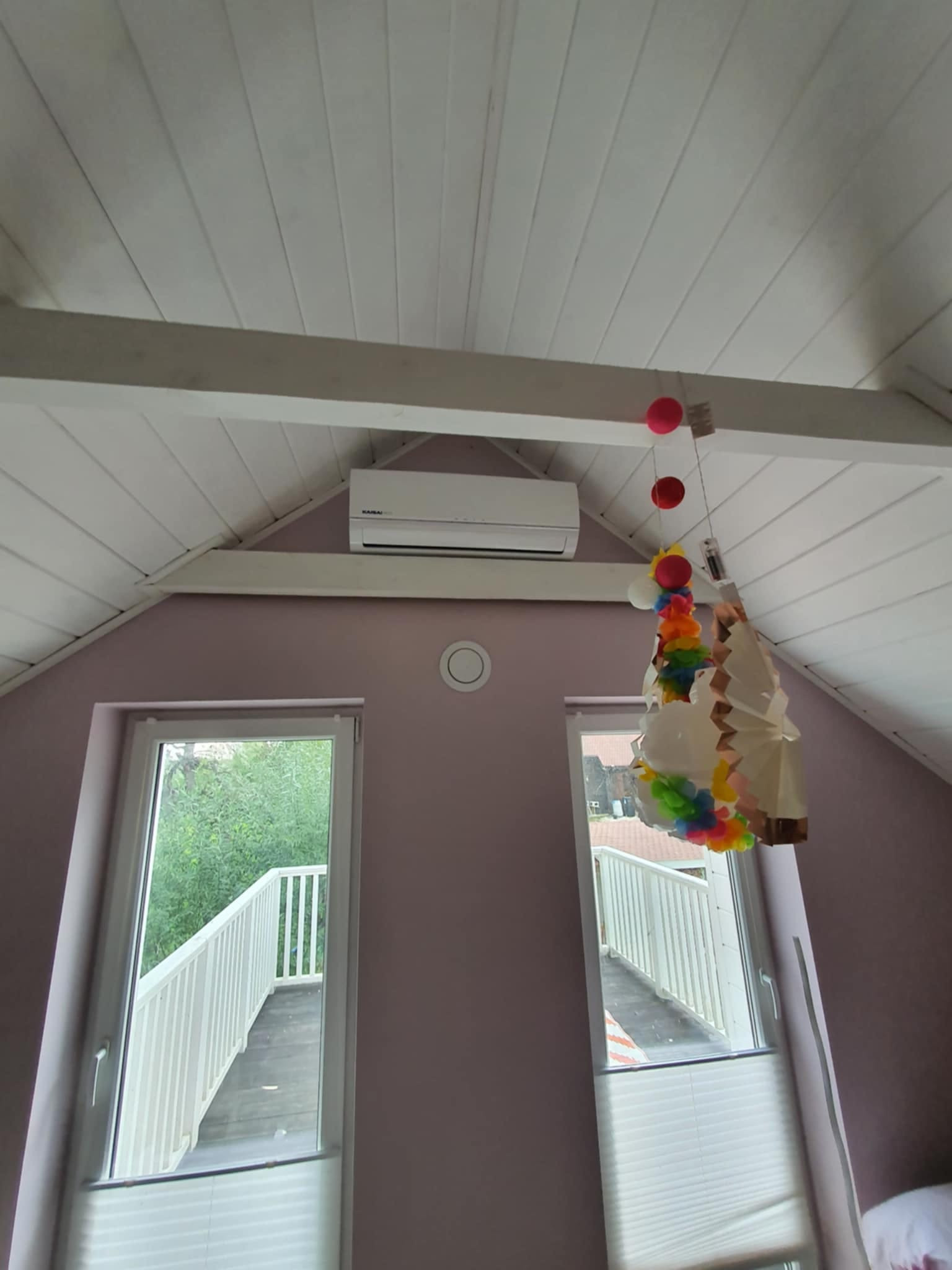 Biały klimatyzator zamontowany pod skośnym, drewnianym sufitem w pokoju na poddaszu, widok z dołu, dekoracja wisząca obok okna balkonowego.