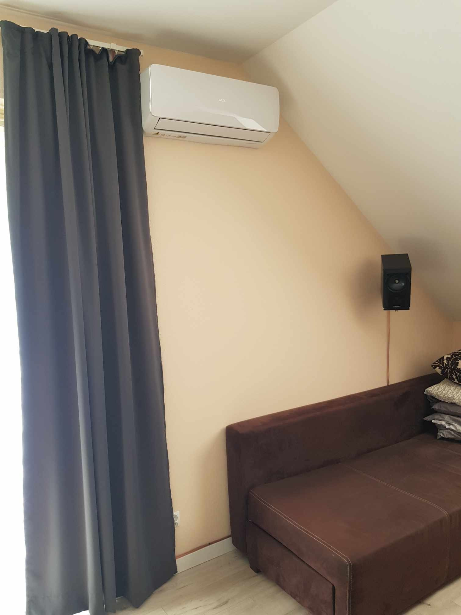 Biały klimatyzator ścienny zamontowany w pokoju na poddaszu, obok zasłoniętego okna i głośnika, brązowa sofa w tle.