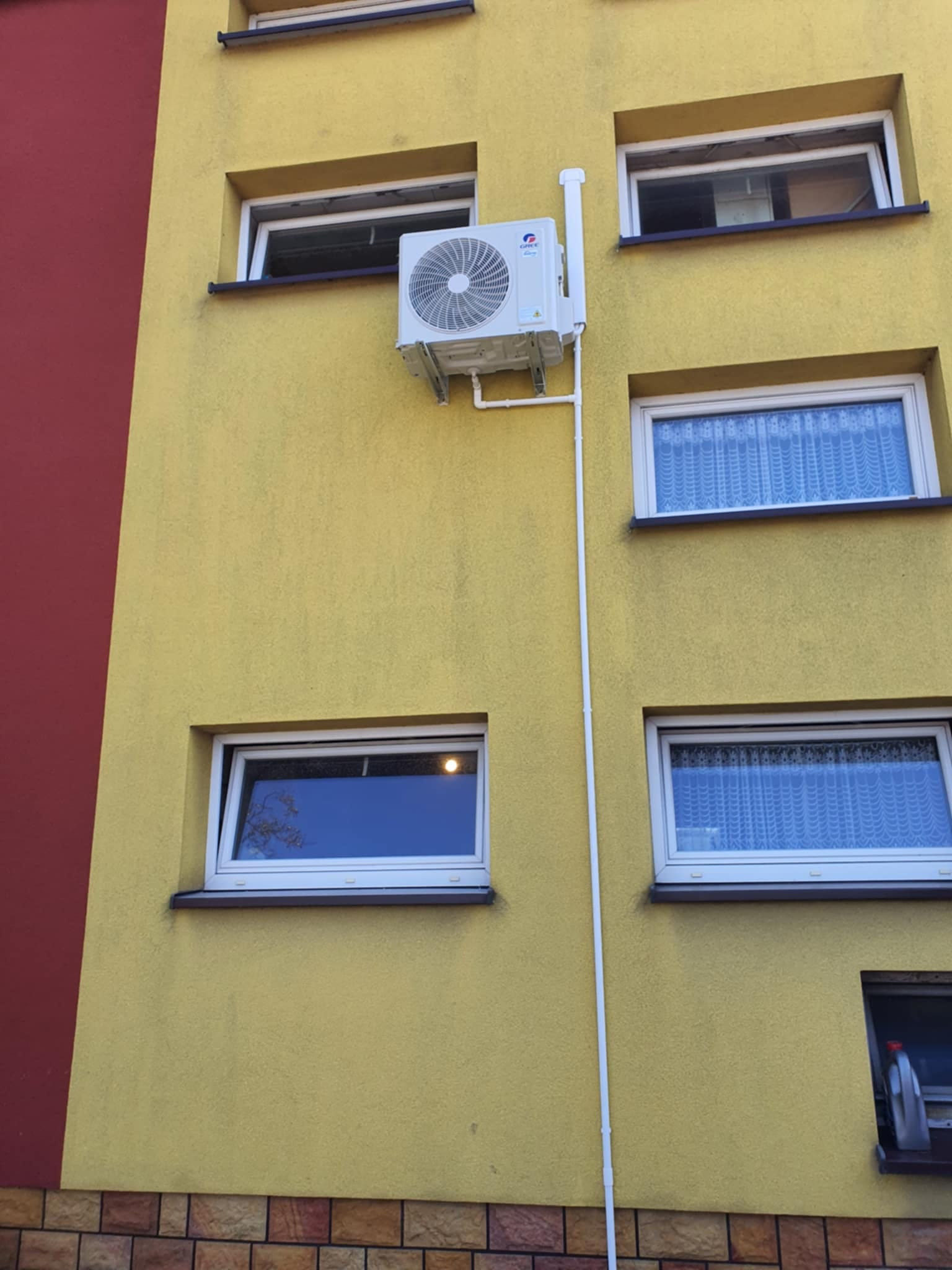 Zewnętrzna jednostka klimatyzatora marki Gree zamontowana na żółtej elewacji budynku mieszkalnego, z widocznymi białymi rurami odprowadzającymi skropliny. W tle czerwony fragment ściany i okna...
