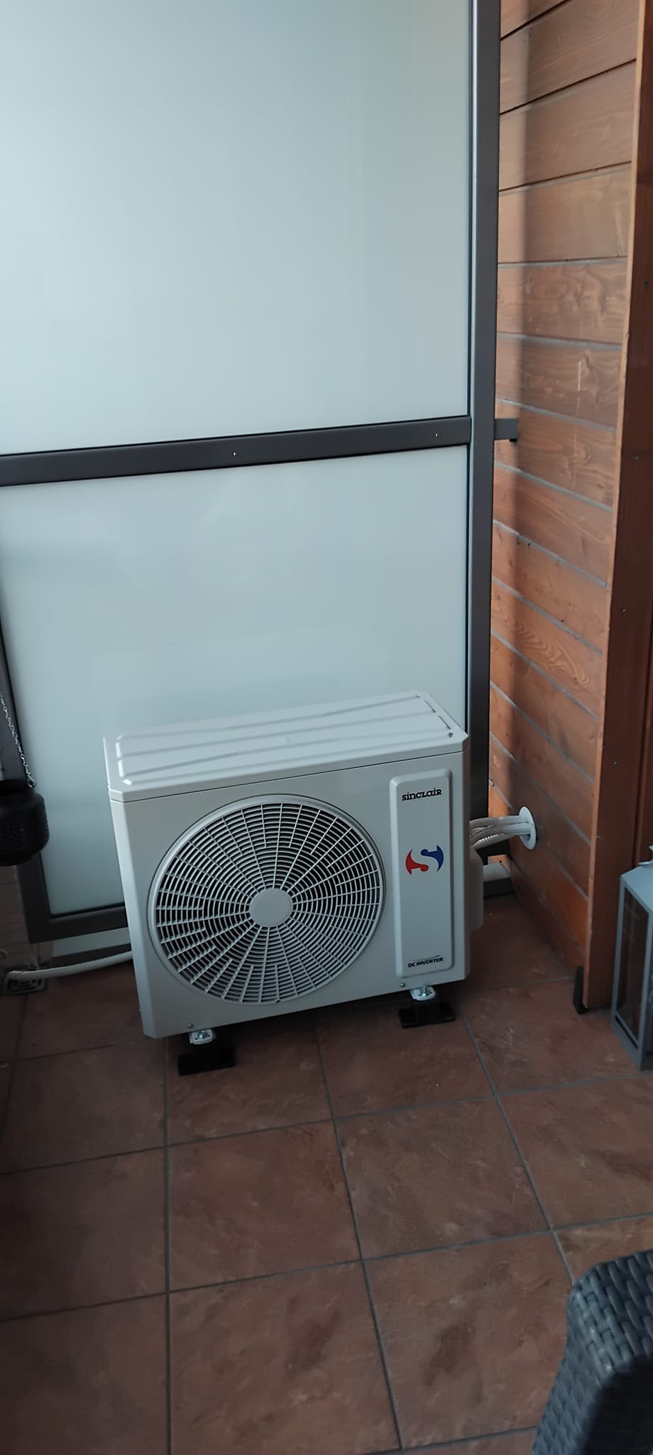 Zewnętrzna jednostka klimatyzacji Sinclair zamontowana na tarasie z brązowymi płytkami i drewnianą ścianą, widoczne przewody wychodzące przez otwór w ścianie.