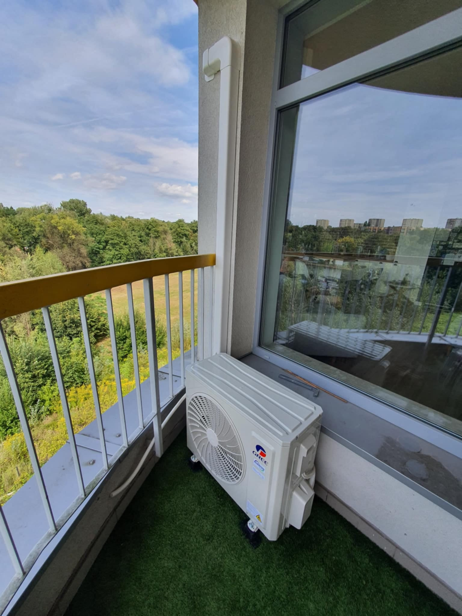 Kompaktowa jednostka zewnętrzna klimatyzatora marki Gree umieszczona na balkonie z imitacją trawy, widok na zieleń i budynki w oddali.