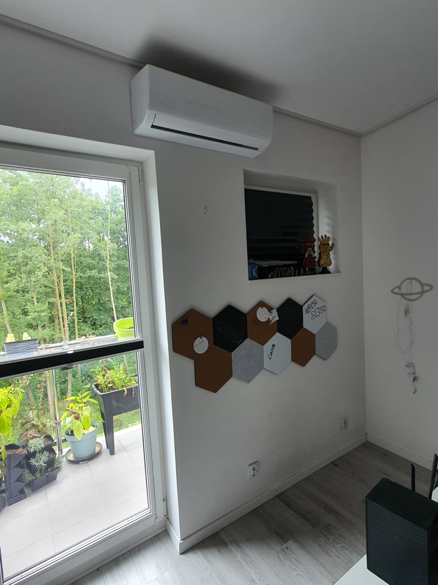 Biały klimatyzator zamontowany na ścianie w nowoczesnym pokoju z widokiem na balkon pełen roślin, dekoracyjna tablica korkowa w kształcie plastrów miodu i małe okno z figurkami.