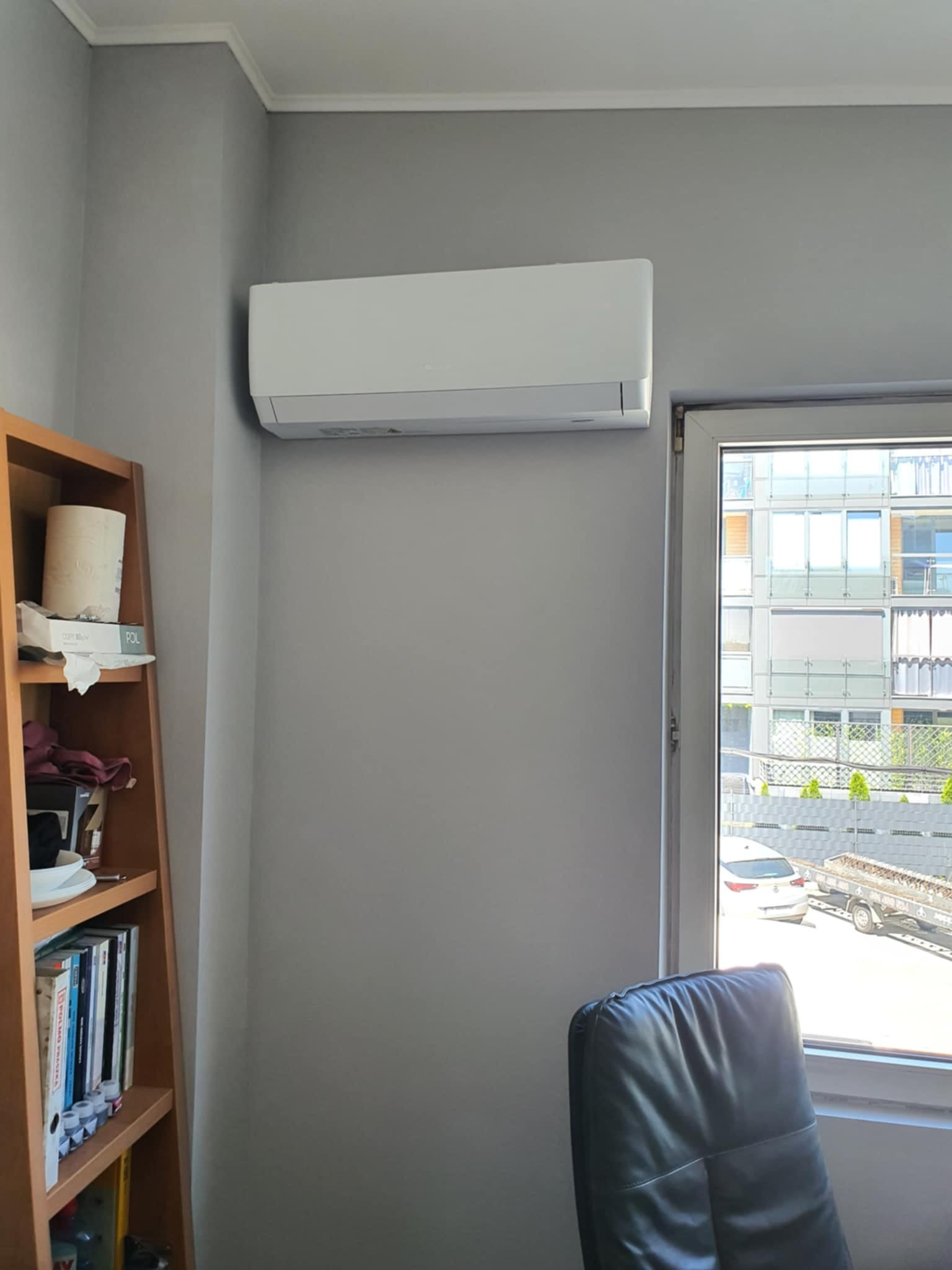 Biały klimatyzator ścienny zamontowany w szarym pokoju, widok zza czarnego skórzanego fotela, za oknem budynek mieszkalny i zaparkowane samochody.