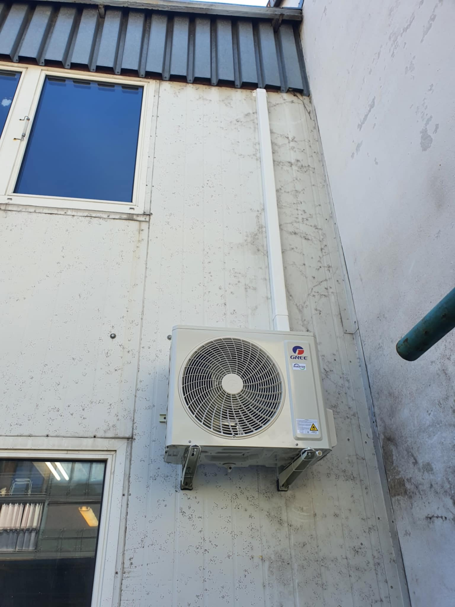 Zewnętrzna jednostka klimatyzacji marki Gree zamontowana na ścianie budynku z widocznymi oknami i rurą spustową.