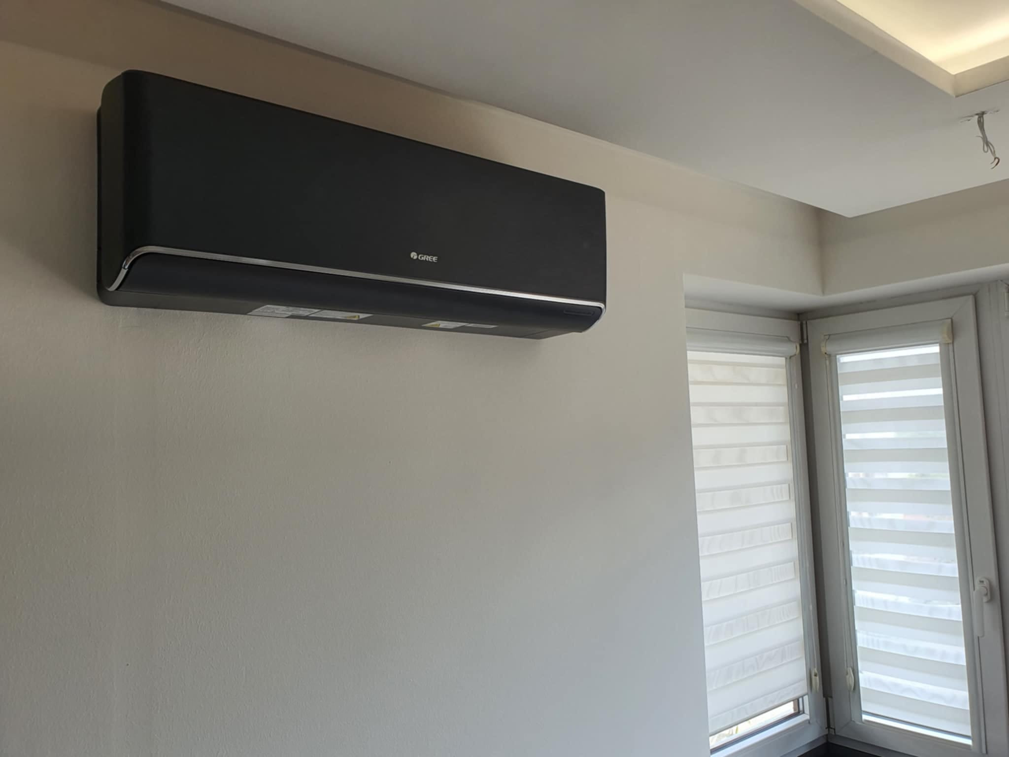 Czarny klimatyzator marki Gree zamontowany na ścianie w jasnym pomieszczeniu z oknem z żaluzjami.