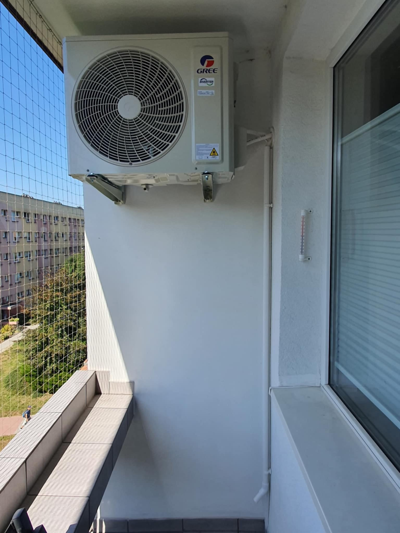 Biała jednostka zewnętrzna klimatyzatora marki Gree zamontowana na balkonie mieszkania w bloku, widok z boku, na ścianie widoczny termometr.
