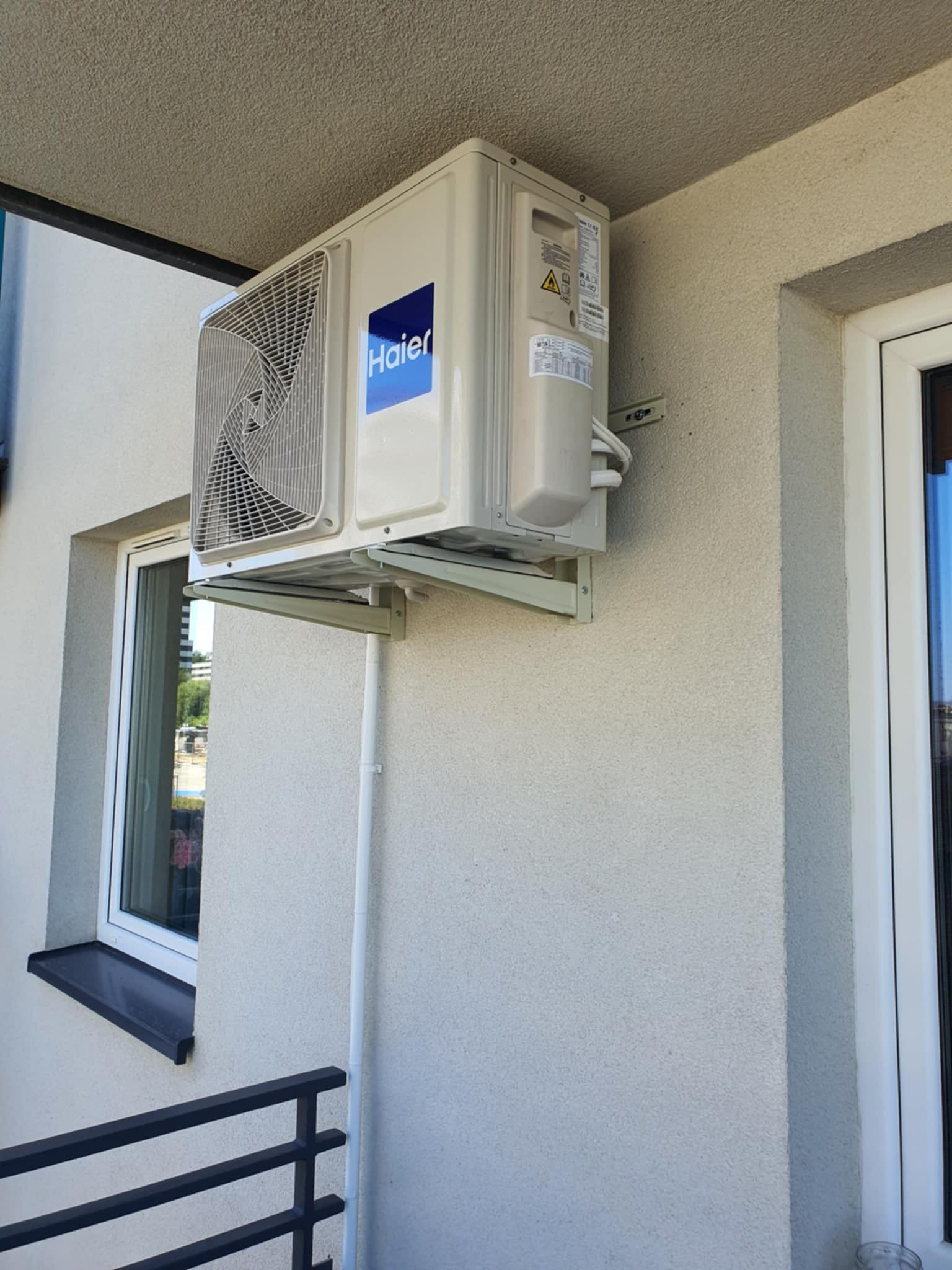 Zewnętrzna jednostka klimatyzacji marki Haier zamontowana na balkonie budynku mieszkalnego, widoczna rura odprowadzająca skropliny.