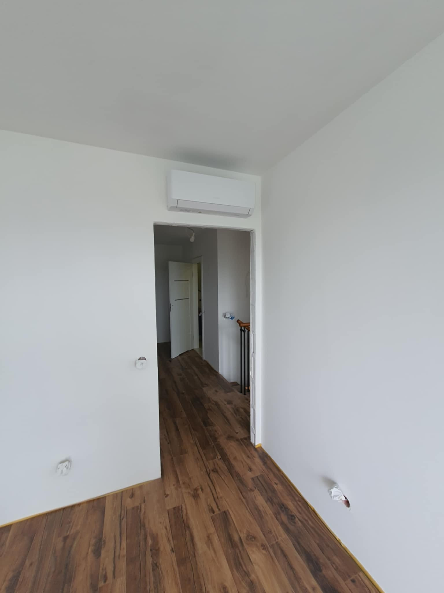 Białe wnętrze z zamontowaną klimatyzacją ścienną, widoczny parkiet i otwarte drzwi do kolejnego pomieszczenia.