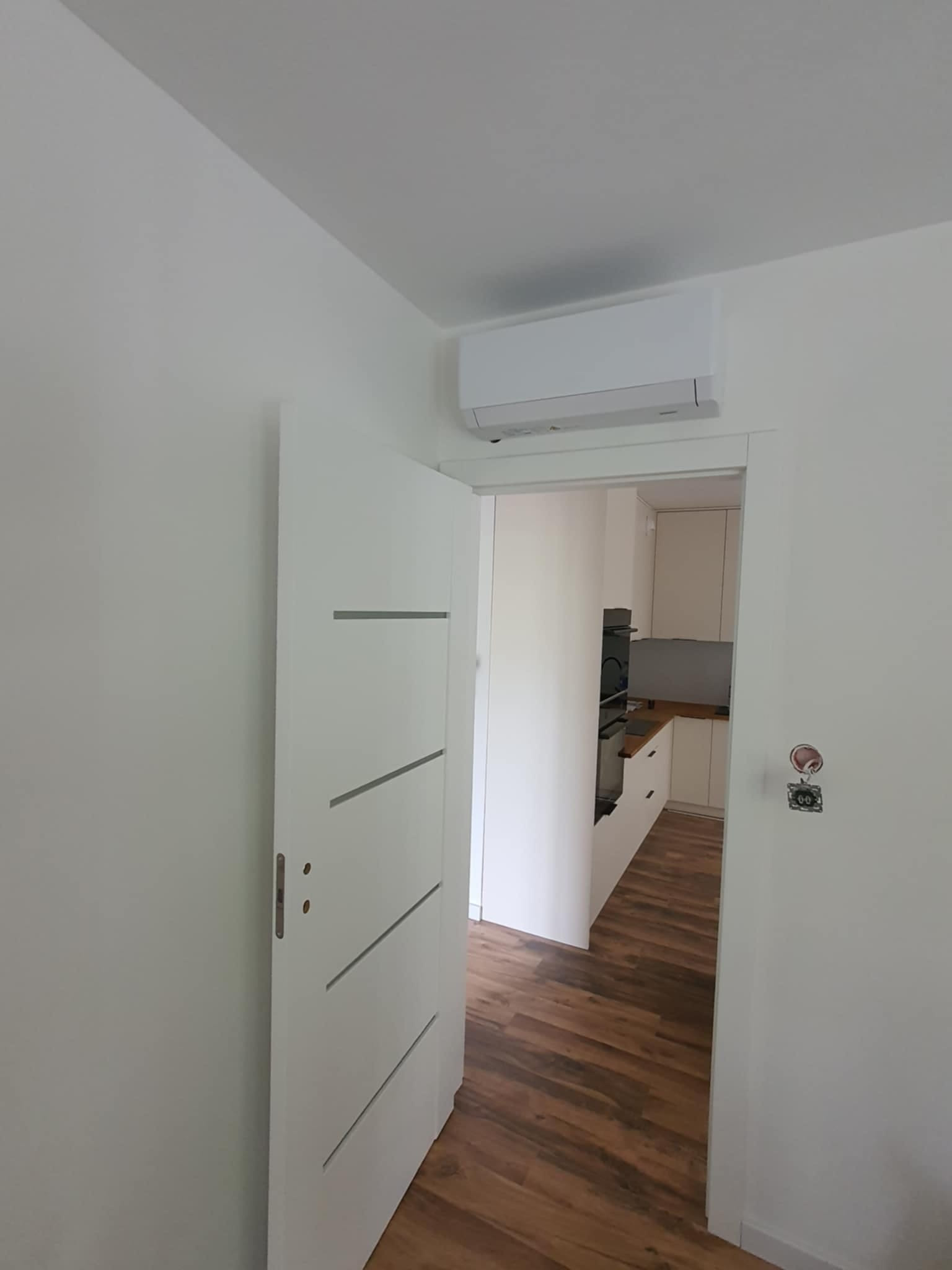 Biały klimatyzator zamontowany na ścianie w jasnym wnętrzu, widok z korytarza na otwartą kuchnię z drewnianą podłogą i białymi meblami.