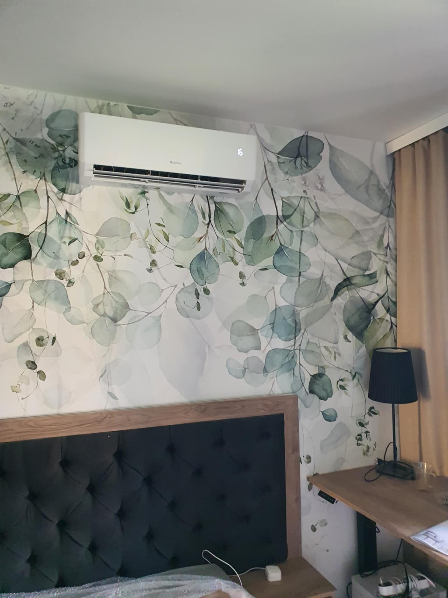 Biały klimatyzator zamontowany na ścianie z motywem liści eukaliptusa w sypialni nad wezgłowiem łóżka.