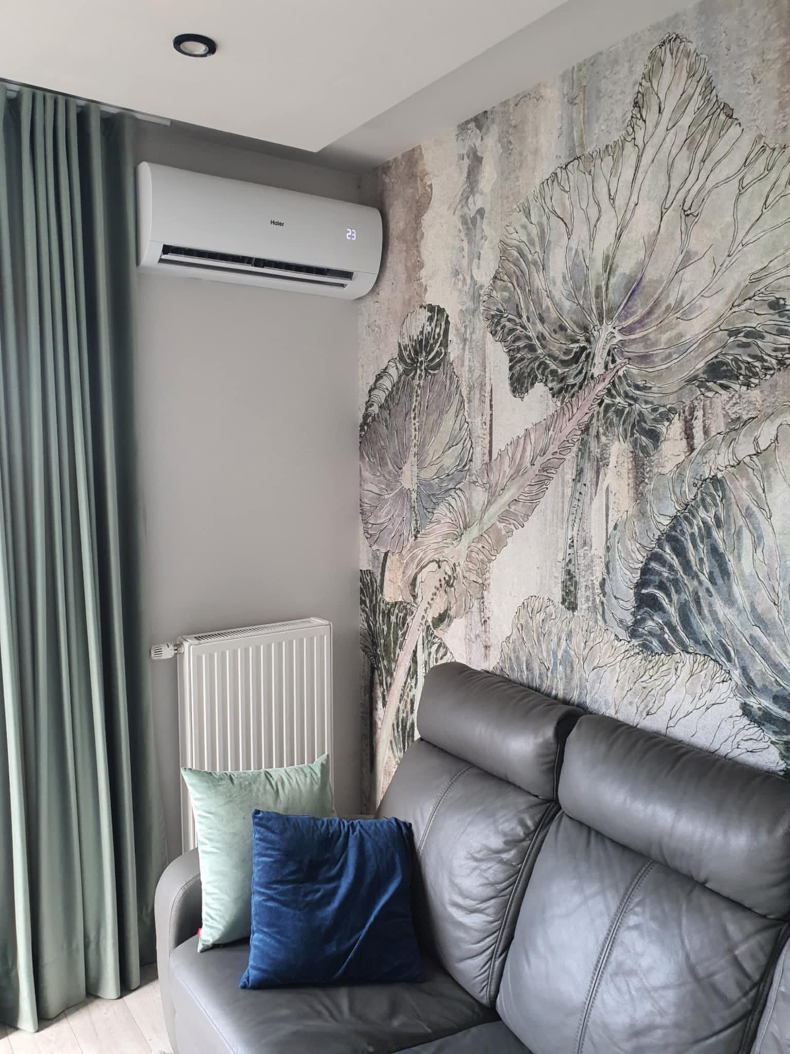 Biały klimatyzator Haier zamontowany na szarej ścianie nad skórzaną kanapą, obok dekoracyjnej tapety z motywem roślinnym i zasłon w kolorze pistacjowym.