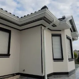 Elewacja budynku po renowacji z oknami z żaluzjami, ozdobnymi gzymsami i rynnami, widok z dołu na tle pochmurnego nieba.