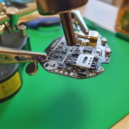 Płytka drukowana smartfona mocowana w uchwycie podczas lutowania elementu, widoczne narzędzia i zielona mata antystatyczna.