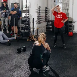 Trening grupowy w siłowni: kobieta w szarej koszulce wykonuje ćwiczenie w podporze, druga w przysiadzie tyłem do kamery, trenerka w czerwonej koszulce obserwuje, w tle mężczyźni i sprzęt do ćwiczeń.