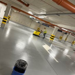 iClean Spółka z ograniczoną odpowiedzialnością - Maszynowe czyszczenie szarej posadzki w garażu podziemnym. Pracownik w żółtej kamizelce obsługuje szorowarkę. W tle słupy z czarno-żółtymi pasami i instalacje pod sufitem.