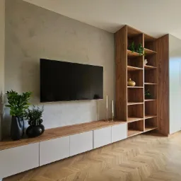 Nowoczesny salon z szafką RTV na wymiar, drewnianym regałem i dekoracyjnym tynkiem na ścianie. Parkiet w jodełkę i rośliny doniczkowe dodają wnętrzu przytulności.