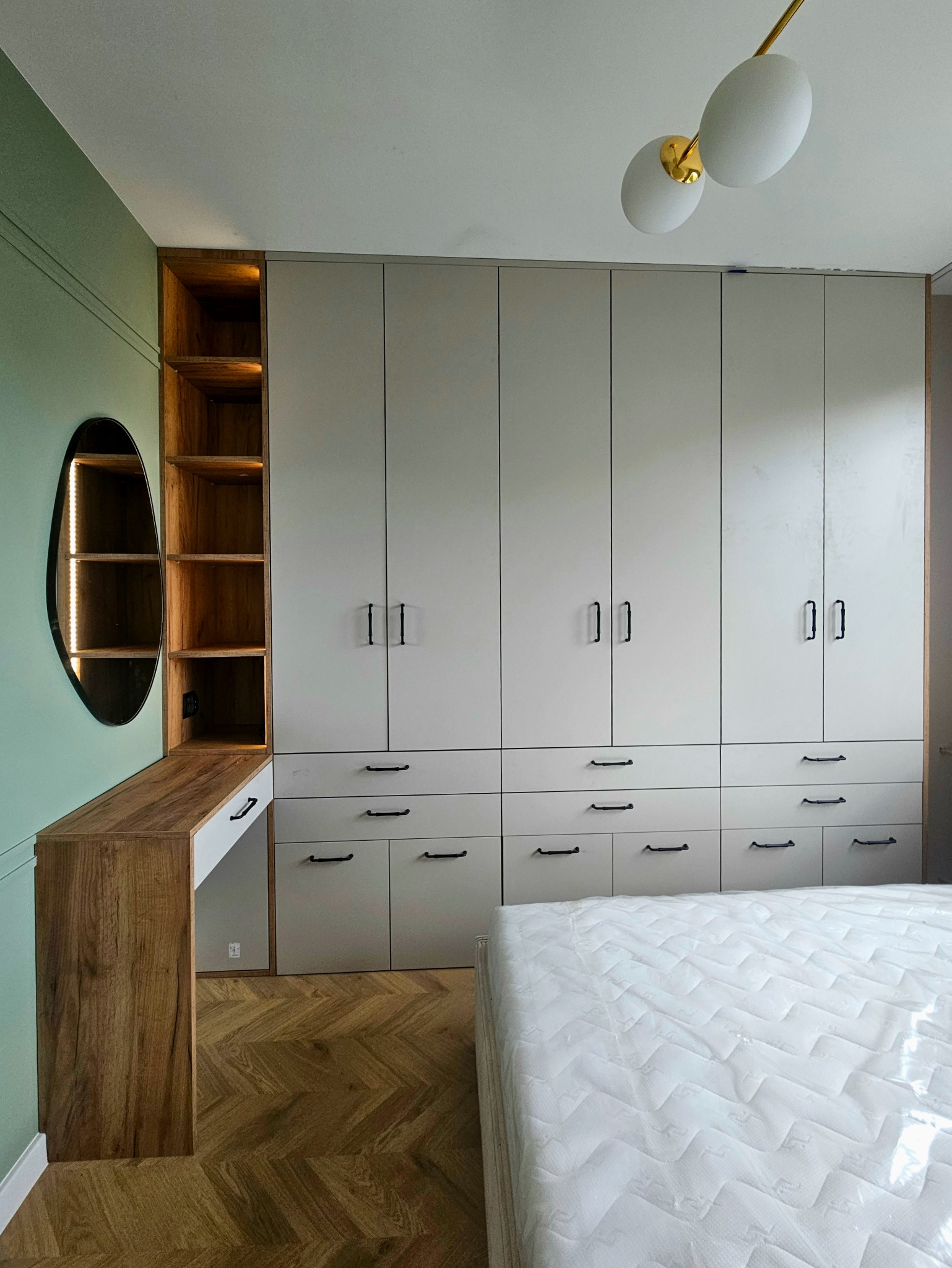 Szafa wnękowa z szufladami i półkami, drewniany blat z owalnym lustrem, parkiet w jodełkę, lampa sufitowa. Minimalistyczny design szafy w sypialni.