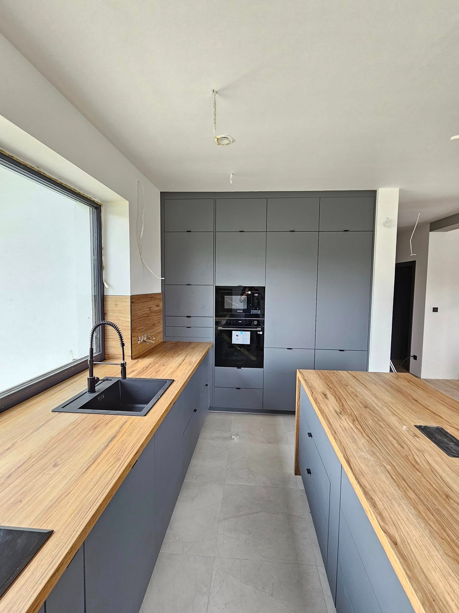 Szara kuchnia z drewnianym blatem i wyspą, czarny zlew z baterią sprężynową, zabudowa z piekarnikiem i mikrofalą, surowy stan wykończenia.