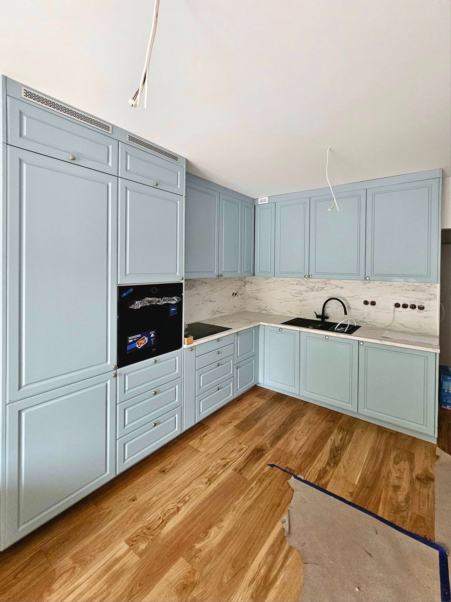 Jasna, narożna zabudowa kuchenna z marmurowym blatem i drewnianą podłogą. Widoczne niedokończone prace wykończeniowe.