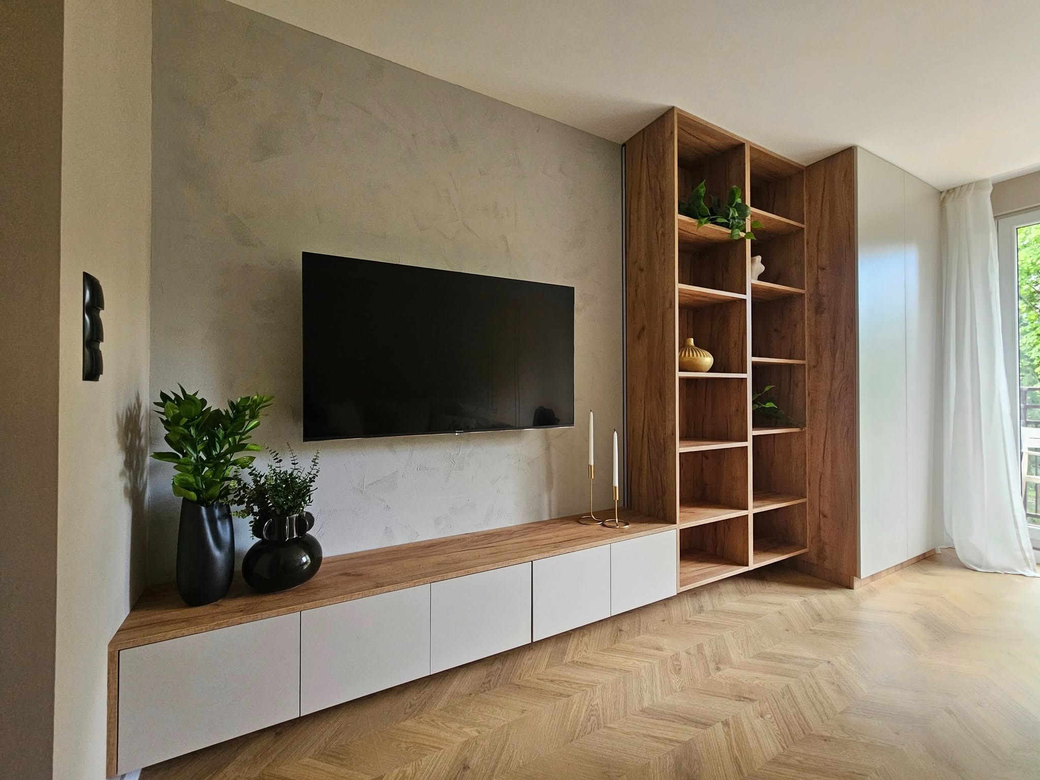 Nowoczesny salon z szafką RTV na wymiar, drewnianym regałem i dekoracyjnym tynkiem na ścianie. Parkiet w jodełkę i rośliny doniczkowe dodają wnętrzu przytulności.