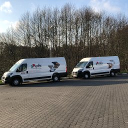 Ruby Studio Spółka z ograniczoną odpowiedzialnością - Dwa białe samochody dostawcze z logo firmy Elis zaparkowane na brukowanym parkingu, z drzewami bez liści w tle i błękitnym niebem.