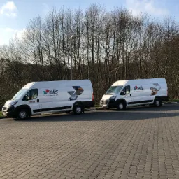 Dwa białe samochody dostawcze z logo firmy Elis zaparkowane na brukowanym parkingu, z drzewami bez liści w tle i błękitnym niebem.