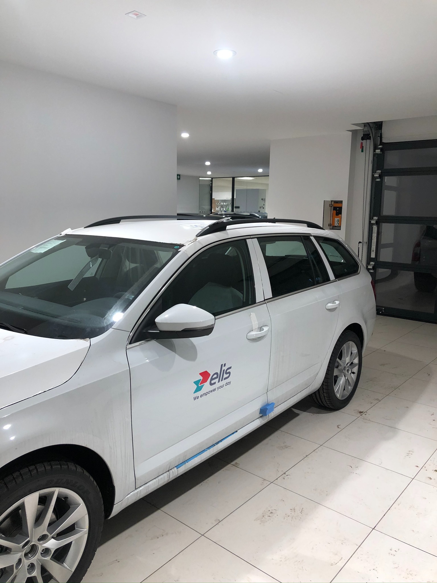 Biały samochód typu kombi z logo firmy 'elis' na boku, przygotowywany do oklejania, z taśmą maskującą i gąbkami zabezpieczającymi, stojący w czystym garażu z białymi ścianami i podłogą w kratkę.